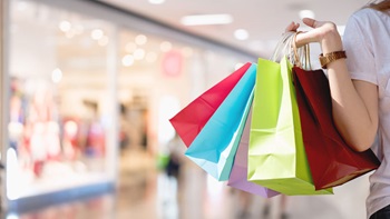 imagem de um braço que segura um conjunto de sacos de compras coloridos, com montra de loja ao fundo